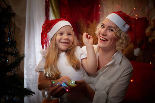 Cute mother and daughter in the red hats of Santa Claus assistants in a room decorated for Christmas. The tradition of decorating house and dressing up for holidays. Happy childhood and motherhood