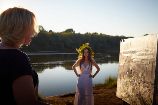 Adult female artist painting picture near water of river or lake in nature. Girl in white sundress and flower wreath posing for holiday of Ivan Kupala in nature at sunset near artist