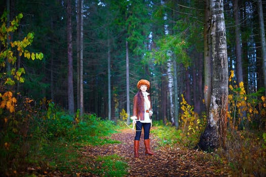 Girl in a leather jacket, a big red fox fur hat and with a crossbow in the forest in autumn. A female model poses as a fabulous royal huntress on nature hunt at a photo shoot
