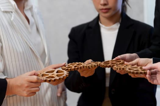 Closeup hand holding wooden gear by businesspeople wearing suit for harmony synergy in office workplace concept. Group of people hand making chain of gears into collective form for unity symbol.