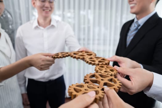 Closeup hand holding wooden gear by businesspeople wearing suit for harmony synergy in office workplace concept. Group of people hand making chain of gears into collective form for unity symbol.