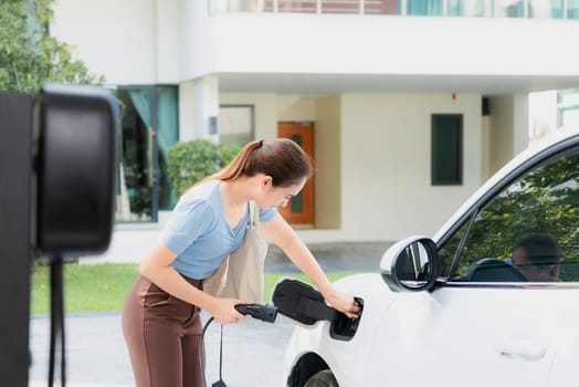 Progressive woman install cable plug to her electric car with home charging station. Concept of the use of electric vehicles in a progressive lifestyle contributes to clean environment.