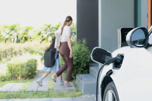 Focus electric vehicle recharging at home charging station plugged in with EV charger device with blurred background of progressive mother and daughter walking as concept for sustainability of energy.