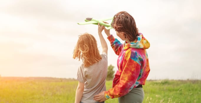 Mother and daughter launching toy plane at the field at summer. Girl and woman spending family time together at the nature. Portrait from back with sunset rays