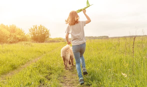 Preteen child girl running with golden retriever dog in the field and holding toy airplane in her hands. Kid with pet doggy playing with plane at the nature