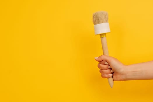 Woman hand holding wooden paint brush up on yellow background with copy space. Concept of creativity and inspiration. Working tool for home design project