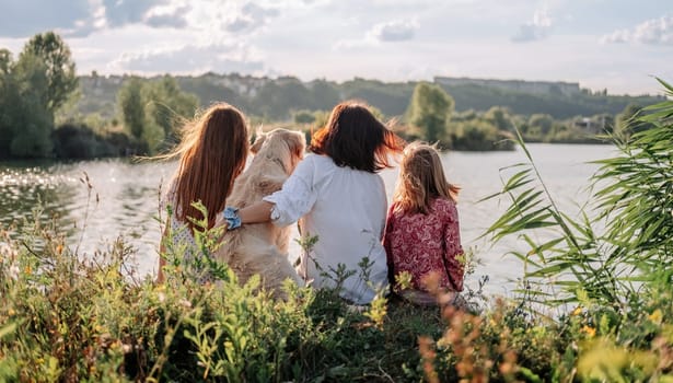 Family with golden retriever dog sitting outdoors at the nature and looking at the lake. Mother with daughters and pet doggy resting close to river