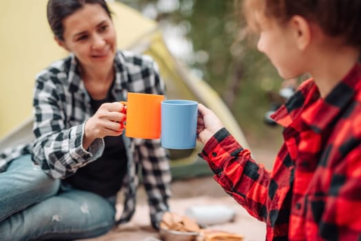 Beautiful woman with girl drinking tea in camping in forest with tent on background and smiling. Young mother and her teenager daughter resting with picnic in the wood