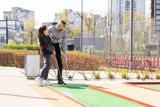 Sharing with golf experience. Cheerful young man teaching his daughter to play mini golf at the day time. Concept of friendly family.