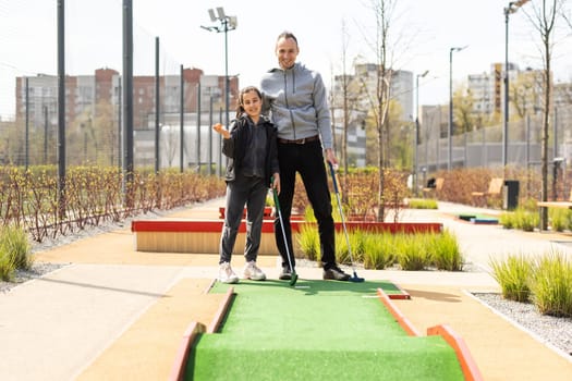Sharing with golf experience. Cheerful young man teaching his daughter to play mini golf at the day time. Concept of friendly family.