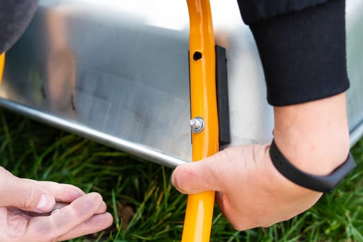 A metal wheelbarrow stands on the street, a man twists a new wheelbarrow for work, twists bolts and nuts