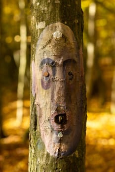 A sad human face in the form of an icon is painted on the trunk of a tree. Defocused bokeh background, all in pastel shades, in a forest full of day light