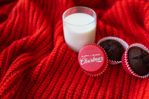 Chocolate cupcakes lie together with a glass of milk on a red knitted plaid, the inscription on the form for cupcakes happy and merry christmas