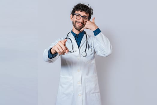 Smiling doctor making call gesture and pointing at the camera. Young doctor making call gesture isolated