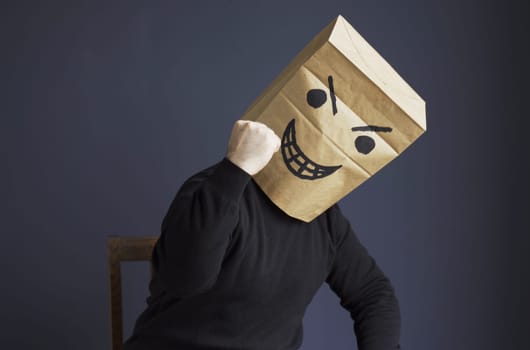 A man in a black turtleneck with a bag on his head, with a drawn angry emoticon, shows his fist while sitting on a chair. Emotions and gestures.
