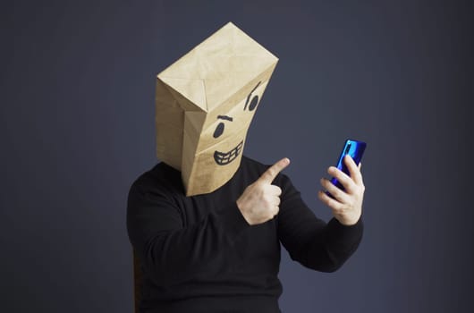 A man in a black turtleneck with a paper bag with an angry emoticon on his head is talking on a video link on a smartphone, gesturing with his hand. Emotions and anger