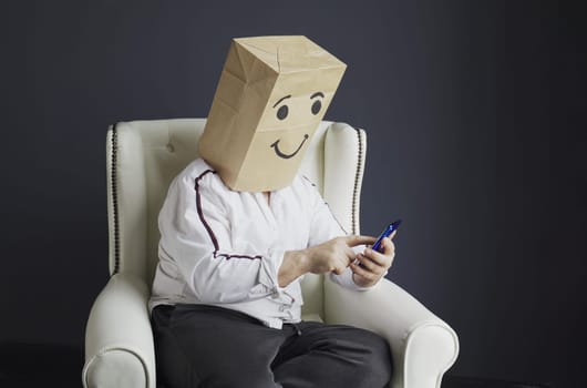 A man in a white shirt with a paper bag on his head, with a smiley face drawn, is typing a message on a smartphone.