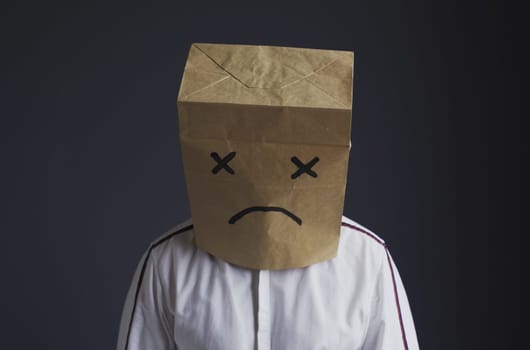 A man in a white shirt with a bag on his head, with a sad emoticon drawn, stands with his head down.