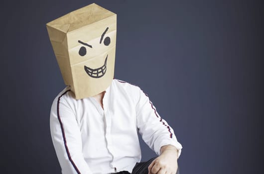 A man sits on a chair with a paper bag on his head, with a drawn angry smiley. Emotions and gestures.