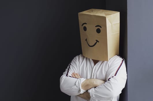A portrait of a man in a white shirt with a paper bag on his head, with a painted smiley face, stands against the wall.