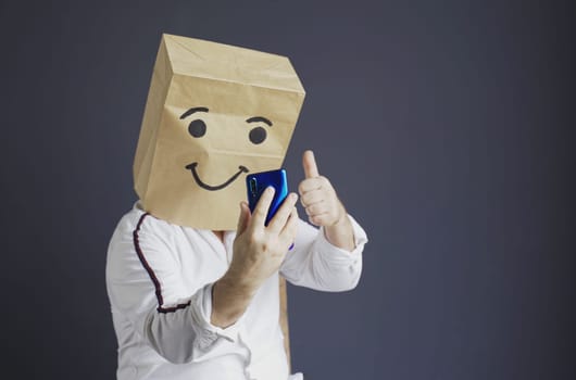 A man in a white shirt with a paper bag on his head, with a drawn smiley face, is talking on a video link or taking a selfie on a smartphone.