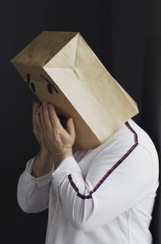 A sad man in a white shirt with a bag on his head, with a drawn crying emoticon, stands against the wall and cries. Emotions and gestures. Vertical frame.