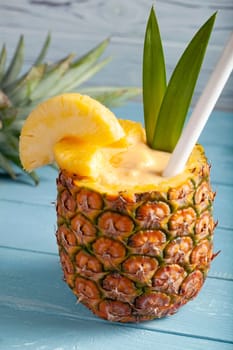 A vertical photo of a pina colada cocktail on a blue wooden background with pineapple pieces, a white straw, two green leaves, and a sliced pineapple crown lying in the blurred background