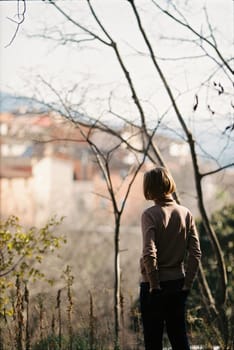 A 12 year old teenage boy in a sweater stands with his back to the camera on a high hill in a small forest and looks into the distance at a small town