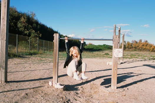 Playful little girl in leather jacket playing on sports ground while hanging on bar during weekend sunny day