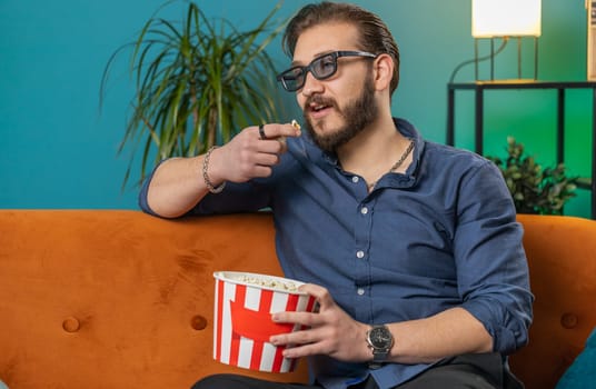 Excited lebanese man in 3D glasses sits on couch eating popcorn snacks, watching interesting TV serial sport game, film online social media movie content at home apartment. Guy enjoying entertainment