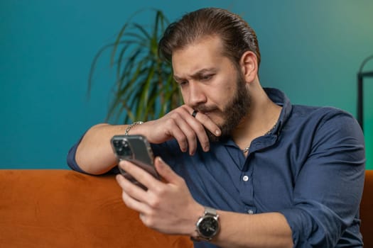 Sad displeased lebanese man use smartphone typing browsing, loses becoming surprised sudden lottery results, bad news, fortune loss, fail, deadline, virus. Young arabian guy at home room on couch