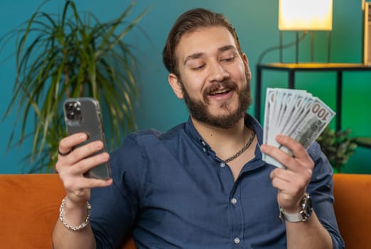 Planning family budget. Smiling lebanese man counting money cash use smartphone calculate domestic bills at home room. Joyful arabian guy satisfied of income and saves money for planned vacation gifts