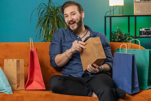 Young lebanese man happy shopaholic consumer came back home after shopping sale with bags. Portrait of arabian guy satisfied received parcels purchase from online order at home apartment room on sofa