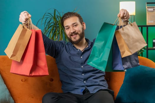 Young lebanese man happy shopaholic consumer came back home after shopping sale with bags. Portrait of arabian guy satisfied received parcels purchase from online order at home apartment room on sofa