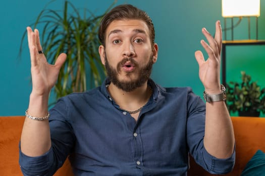 Oh my God, Wow. Young lebanese man surprised looking at camera with big eyes shocked by sudden victory good win news, celebrating. Portrait of excited amazed arabian guy at home apartment room on sofa