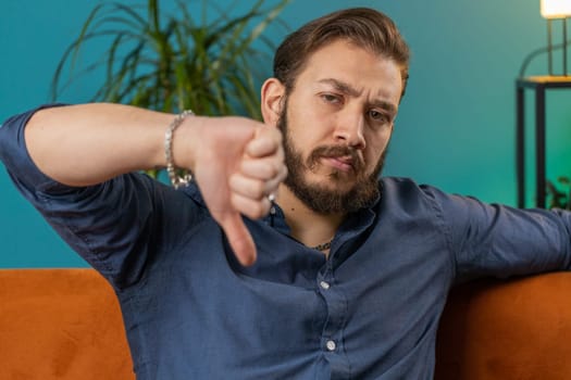 Dislike. Upset lebanese man showing thumbs down sign gesture, expressing discontent, disapproval dissatisfied bad work at modern home apartment indoors. Displeased arabian guy in living room on sofa
