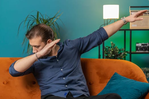 Trendy cheerful positive young lebanese man having fun dancing and moving to rhythm dabbing raising hands making dub dance gesture. Portrait of arabian guy at modern home apartment living room on sofa