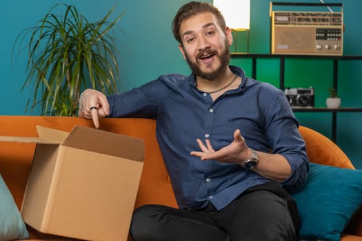 Happy lebanese man unpacking delivery parcel sitting at home apartment. Smiling satisfied arabian guy shopper online shop customer opening cardboard box receiving purchase gift by fast postal shipping