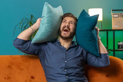 Repair work at neighbours. Irritated mad man relaxing on couch cover ears with pillows annoyed by noisy neighbors suffer from headache wish silence. Thin walls at home flat without sound insulation