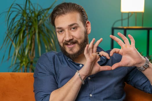 I love you. Happy lebanese man at home living room couch makes symbol of love, showing heart sign to camera, express romantic feelings express sincere positive feelings. Charity, gratitude, donation