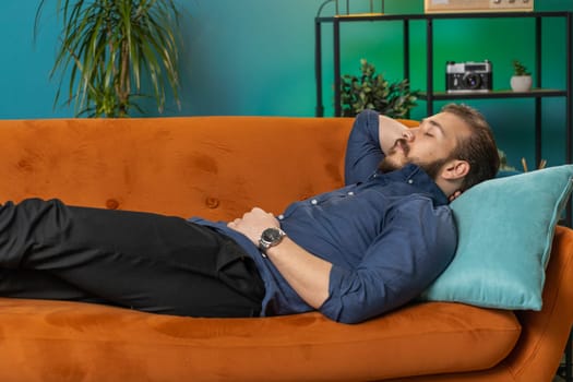 Tired lebanese man lying down on comfortable couch taking a rest, daytime nap. Portrait of carefree arabian guy holding hands behind head relaxing, falling asleep at home apartment living room on sofa