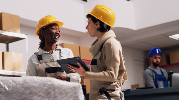 Multiethnic team of women checking stock of goods in warehouse, planning industrial distribution with packages. Two people in overalls and hardhat using digital tablets. Handheld shot.