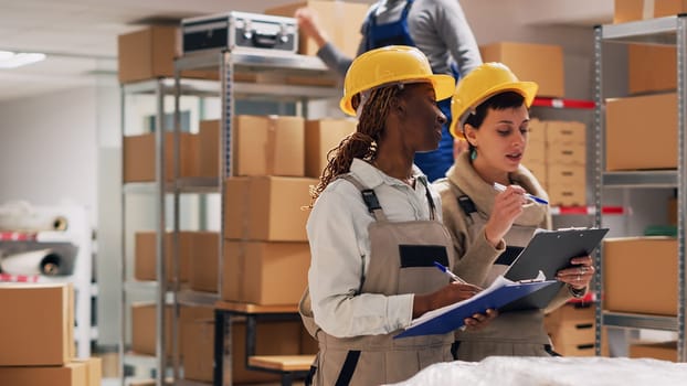 Diverse workers checking inventory on list in depot and counting number of products in cardboard packages. Young people looking at manufacturing goods, industrial merchandise stock.