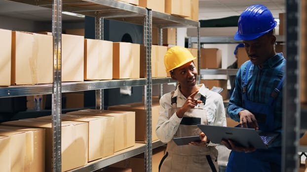 African american people analyzing goods on laptop and tablet, using devices to create stock inventory in storehouse space. Young man and woman working with cardboard packages and cargo.