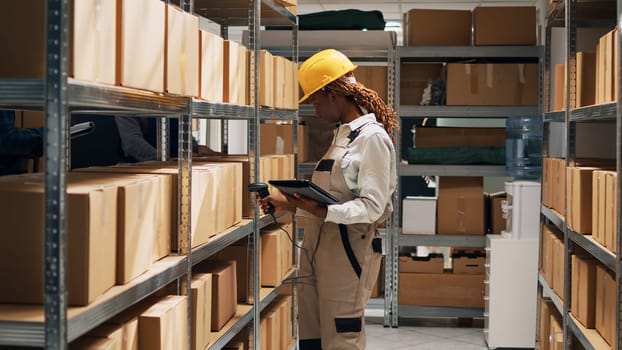 African american woman holding tablet to scan barcodes to work on inventory, using scanner and sorting warehouse merchandise. Depot worker checking cardboard boxes on shelves.