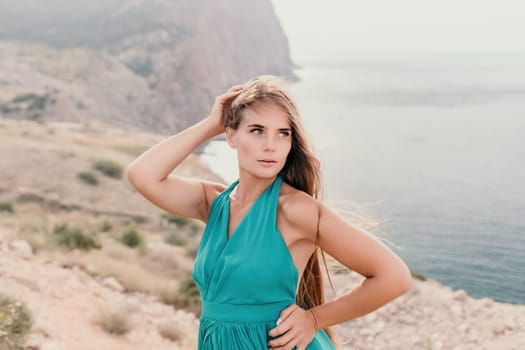 Side view a Young beautiful sensual woman in a mint long dress posing on a volcanic rock high above the sea during sunset. Girl on the nature on overcast sky background. Fashion photo