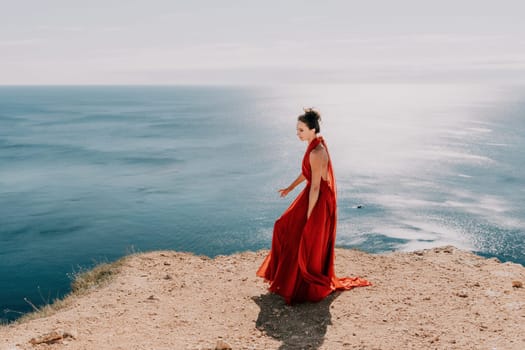 Side view a Young beautiful sensual woman in a red long dress posing on a rock high above the sea during sunrise. Girl on the nature on blue sky background. Fashion photo.