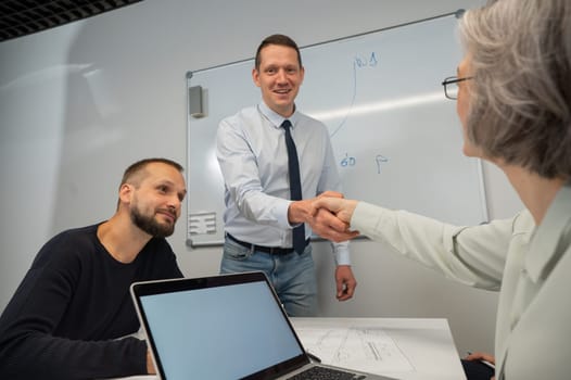 The boss makes a presentation to subordinates at the white board. Caucasian man shaking hands with middle aged woman