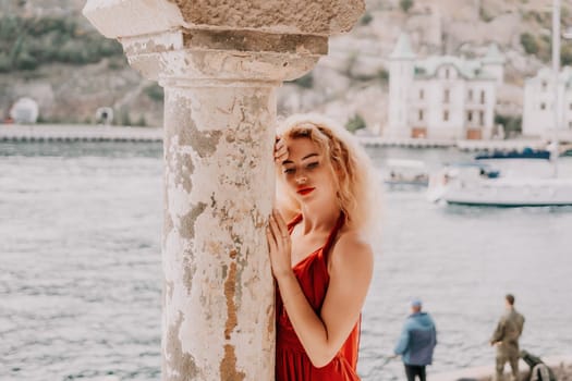 Close up shot of beautiful young caucasian woman with curly blond hair and freckles looking at camera and smiling. Cute woman portrait in a pink long dress posing on a volcanic rock high above the sea