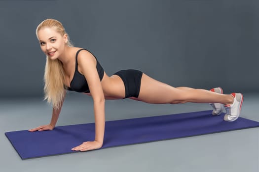 Young beautiful woman in fitness wear trained female body, doing push-ups on the mat. Look at the camera
